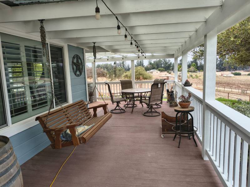 A spacious, freshly painted porch featuring light blue siding, white railings, and a white ceiling with string lights. A wooden porch swing hangs on the left, while an outdoor dining set with a round table and chairs is visible on the right. The porch overlooks a dry, rural landscape with fields and trees under a bright sky.