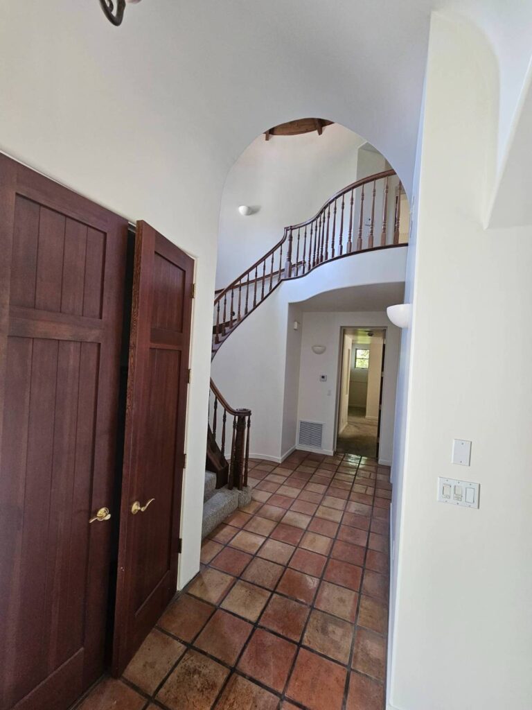A residential interior hallway featuring freshly painted white walls, dark wood bi-fold doors, terracotta tile flooring, and a curved wooden staircase with a banister leading to the upper floor. A doorway at the far end of the hall opens into another room.