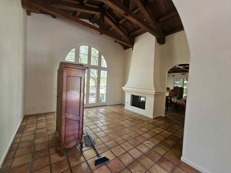 A spacious living room with freshly painted white walls, a high ceiling featuring dark exposed wood beams, and warm terracotta tile flooring. A large arched window with French doors offers a view of outdoor greenery, and a white fireplace is built into the right wall. A tall wooden cabinet stands on the left, with a mop and broom resting on the floor.