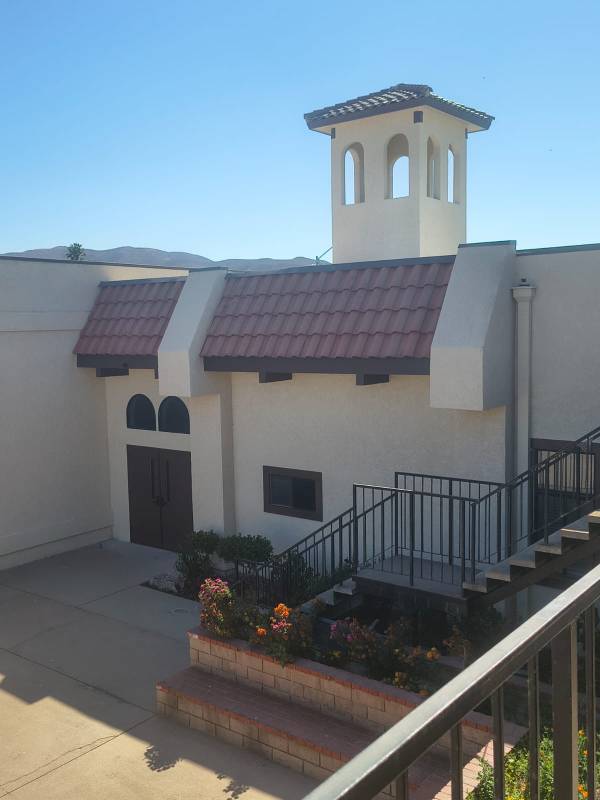 Exterior view of a Spanish-style building painted in light beige stucco with terracotta roof tiles, dark trim, and an architectural tower.