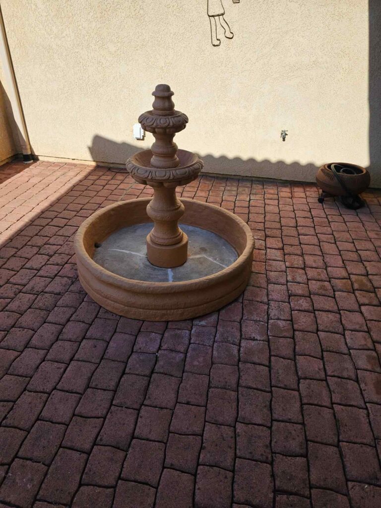 A three-tiered terracotta fountain sits on a red brick paver patio, with a light-colored stucco wall in the background and shadows cast by the sun.