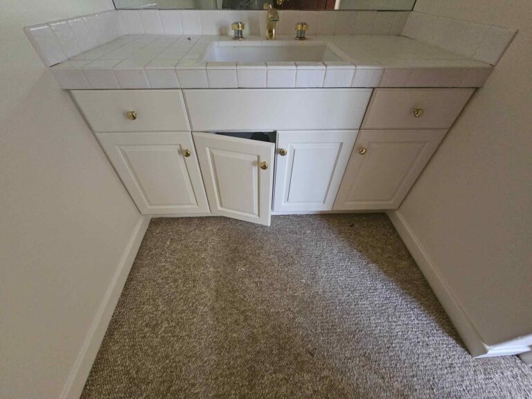 A white bathroom vanity with a tiled countertop and an integrated sink. The vanity features white cabinets with gold knobs, one cabinet door is slightly ajar. The floor in front is carpeted.